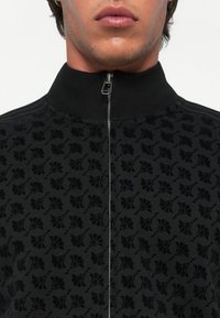 Zwarte zip-up jas met een textuur met bloemmotief, geribbelde kraag en ritsdetails. Het materiaal lijkt zacht en licht pluizig.