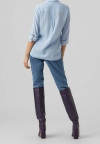 Personne portant une chemise rayée bleue et blanche, un jean bleu et des bottes à talons violettes foncées jusqu'aux genoux, se tenant avec une main sur la hanche.