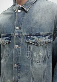 Φωτεινό γαλάζιο τζιν μπουφάν με μεταλλικά κουμπιά, θήκες στο στήθος με καπάκια και διακριτικές φθαρμένες λεπτομέρειες στην δεξιά τσέπη.