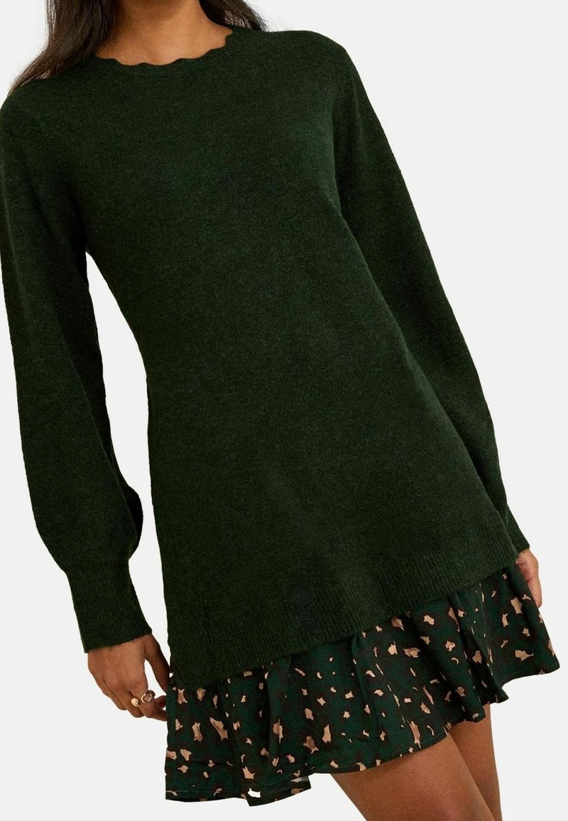 Femme portant une robe-pull à manches longues vert foncé superposée sur une jupe foncée avec des taches abstraites marron clair.