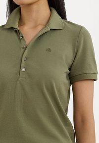 Polo shirt vert olive avec un tissu texturé, patte de boutonnage à trois boutons et un petit logo brodé sur le côté gauche de la poitrine.