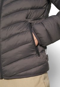 Mørk grå puffjakke med quiltet design, lynlåslommer og elastiske manchetter. Glat, skinnende stof med en subtil tekstur.