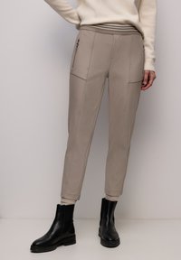 Hellbeige zulaufende Hose mit vorderen Reißverschlusstaschen, getragen mit schwarzen Stiefeletten und cremefarbenem Pullover, gezeigt von der Taille bis zu den Füßen.