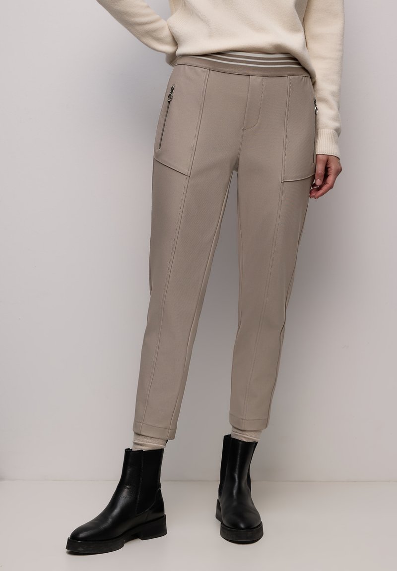 Hellbeige zulaufende Hose mit vorderen Reißverschlusstaschen, getragen mit schwarzen Stiefeletten und cremefarbenem Pullover, gezeigt von der Taille bis zu den Füßen.