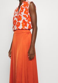 Ärmellose Bluse mit orange-weißen Blumenmuster; kombiniert mit einem fließenden, einfarbigen orangen Rock. Der Stoff wirkt leicht und glatt.