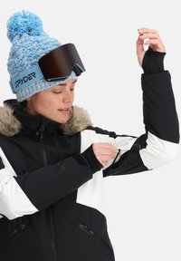 Giacca da sci nera e bianca con colletto in pelliccia, dotata di tasche con cerniera e polsini regolabili. Un berretto a maglia blu e occhiali da sole scuri completano il look.