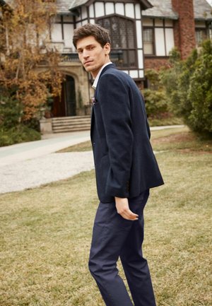 Blazer azul marino con hombros estructurados, tela texturizada y un pañuelo en el bolsillo, combinado con pantalones ajustados azul marino en un entorno exterior con césped.