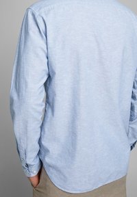 Ljust blå långärmad skjorta i texturerad tyg, med böjd nederkant, knappade ärmslut och en enkel krage.