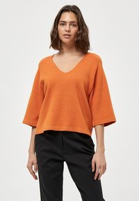 Oranger gestrickter Pullover mit V-Ausschnitt, kurzen weit geschnittenen Ärmeln und einem cropped Saum. Weiche Textur mit glatter Oberfläche. Schwarze Hose darunter getragen.