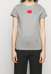 Camiseta de manga corta gris hecha de algodón suave. Cuenta con un parche de logo rectangular rojo con "HUGO" en texto negro en el centro.