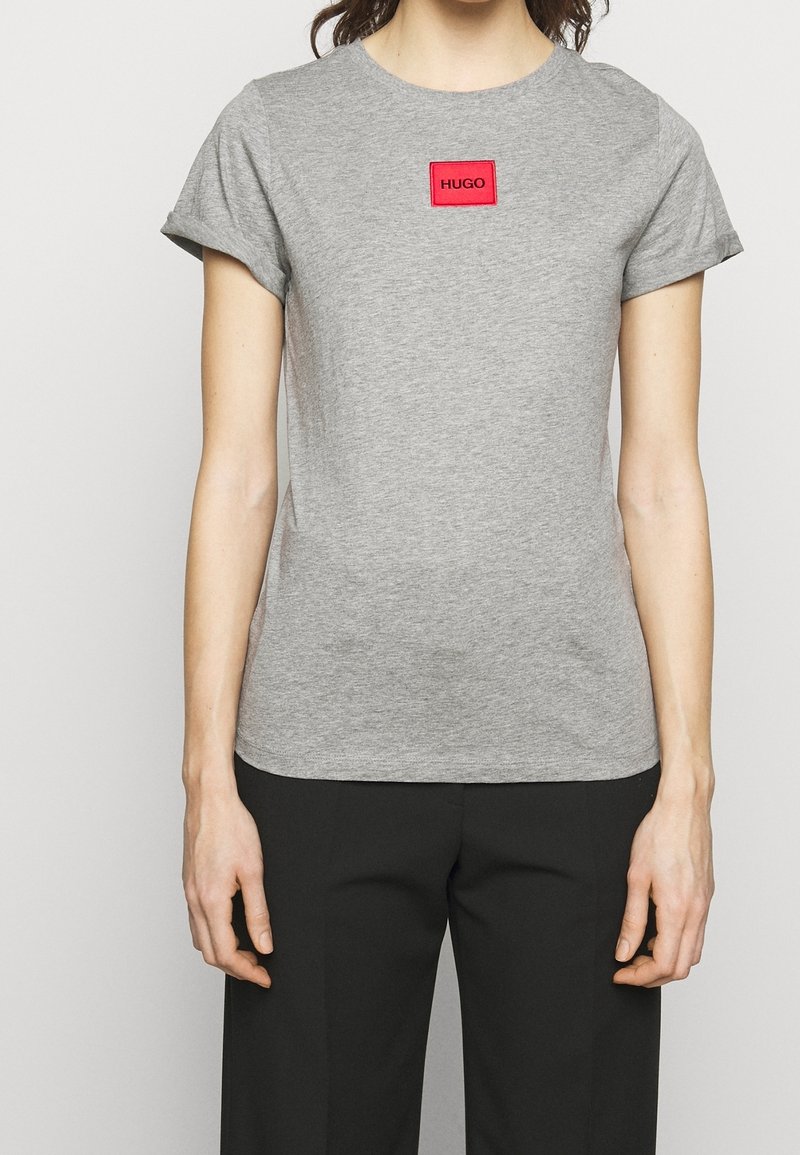 Camiseta de manga corta gris hecha de algodón suave. Cuenta con un parche de logo rectangular rojo con "HUGO" en texto negro en el centro.