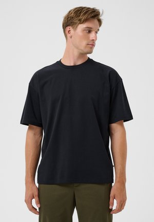 Joven con cabello rubio que lleva una camiseta negra lisa de manga corta y pantalones verde oliva, mirando hacia su derecha sobre un fondo gris.