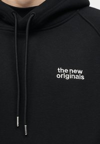 Črna jopa s kapuco in vrvicami, z belim vezenim napisom "the new originals" na prsih.