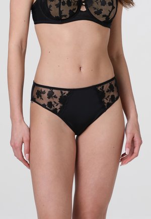 Donna che indossa un reggiseno in pizzo nero con motivo floreale e mutandine abbinate, in piedi davanti a uno sfondo semplice grigio chiaro.