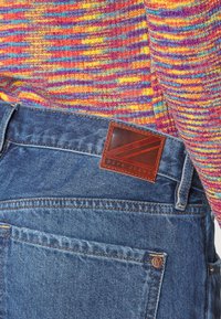 Jeans de mezclilla en azul con un suéter texturizado multicolor encima. Cuenta con una etiqueta de cuero marrón de Pepe Jeans en la parte trasera de la cintura.