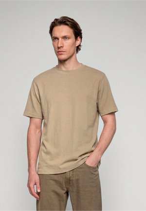 Ung mand med lys hud og brunt hår iført en ensfarvet beige kortærmet t-shirt og brune bukser, den ene hånd i lommen, neutral baggrund.