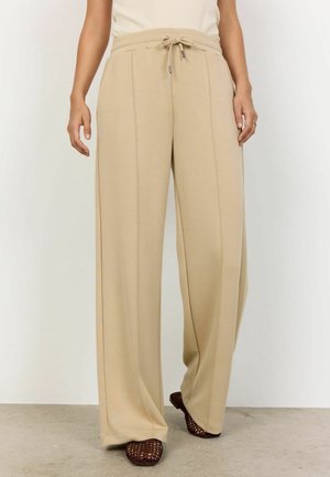 Person wearing beige wide-leg pants with front seam details and a drawstring waist, paired with dark open-knit flat shoes.