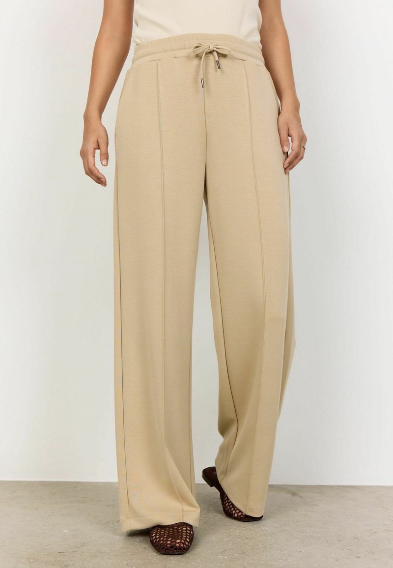 Personne portant un pantalon beige à jambes larges avec des détails de couture à l'avant et une taille à cordon, associé à des chaussures plates en maille ouverte foncées.