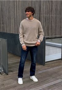 Jonge man die op een houten terras staat, gekleed in een beige gebreide trui, donkerblauwe jeans en witte sneakers, terwijl hij met een lichte glimlach opzij kijkt.