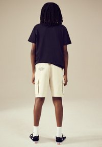 Black short-sleeve t-shirt paired with beige cargo shorts featuring two side pockets and printed text. Black sneakers with white soles and socks.