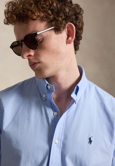 Jeune homme aux cheveux bouclés portant des lunettes de soleil écaille et une chemise bleu clair à boutons avec un petit logo brodé bleu foncé.