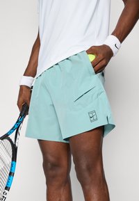 Lyseblå sportshorts med et slankt design, der har en sidelomme og et sort logo på venstre ben. Materialet ser letvægtigt og tekstureret ud.