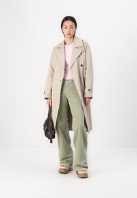 Beige trenchcoat, rosa topp, og lysegrønne vide bukser. Brun teksturert veske og klumpete beige sko med pelsdetaljer. Glatt, tilpasset passform.