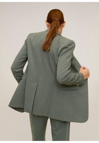 Femme aux cheveux roux en queue de cheval portant un costume léger gris surdimensionné, tenant le revers, de dos contre un fond uni.