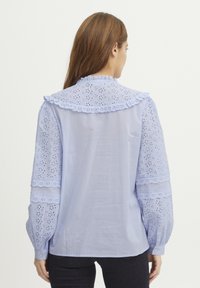 Svetlo modra bluzica z rokavi broderie anglaise, nabranim ovratnikom, nabranimi manšetami in gladkim hrbtom z gumbom za zapiranje.