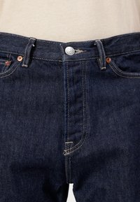 Jean en denim bleu foncé avec un design classique à cinq poches, comportant des rivets en cuivre et des coutures claires. Fermeture à bouton à la taille.