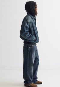 Giubbotto di denim azzurro con colletto classico, indossato sopra un felpa nera, abbinato a jeans blu larghi e scarpe marroni.