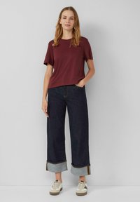 Camiseta corta de mangas en color burdeos combinada con pantalones vaqueros de pierna ancha en un tono oscuro, con los puños enrollados y un tejido gris en contraste debajo. Zapatillas casuales completan el conjunto.