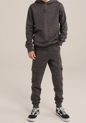 Kind trägt dunkelgrauen Hoodie mit Text und passende Cargo-Jogginghosen, steht mit Händen in den Taschen, schwarz-weiße Sneakers sichtbar.