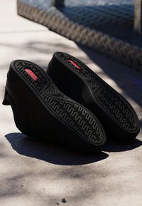 Zapatos negros de cuña con tacón, colocados con la suela hacia arriba sobre una superficie de cemento iluminada por el sol, mostrando patrones detallados de la suela y insertos rojos en la base.
