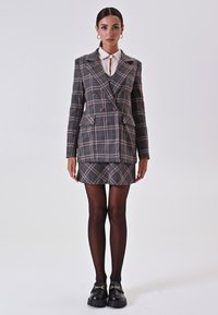 Grau-, Braun- und Rosa-karierter, doppelt geknöpfter Blazer, kombiniert mit einem passenden Minirock. Das Model trägt schwarze Strumpfhosen und klobige schwarze Loafers.