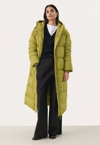 Lange pufferjas in felgroen met een capuchon, gewatteerd ontwerp, drukknoopsluitingen en een ontspannen pasvorm, gecombineerd met zwarte broek en een zwarte cardigan.