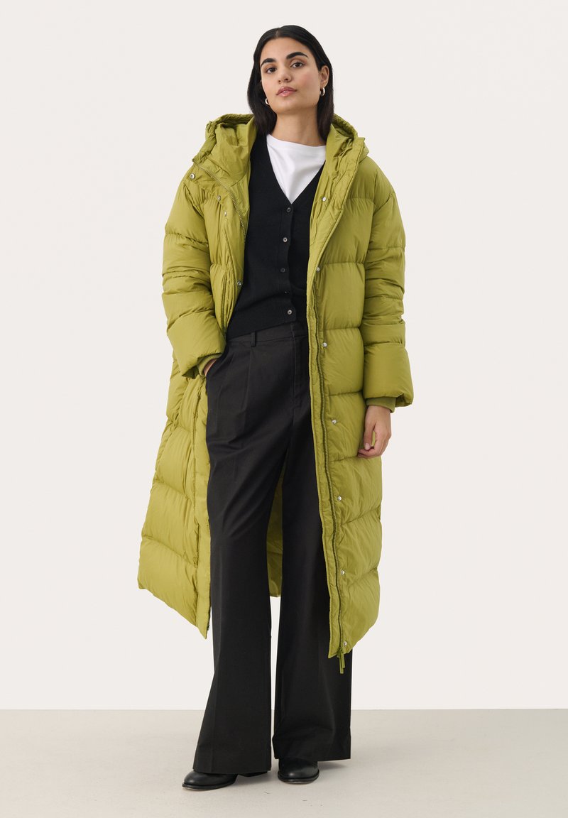 Lange pufferjas in felgroen met een capuchon, gewatteerd ontwerp, drukknoopsluitingen en een ontspannen pasvorm, gecombineerd met zwarte broek en een zwarte cardigan.