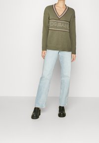 Olivově zelený oversize svetr s V-výstřihem, zdobený logem a páskovými akcenty, kombinovaný se světle modrými nohavicemi rovného střihu a černými botami.