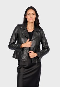 Černá kožená motorkářská bunda s přiléhavým střihem, stříbrným kováním, asymetrickým zapínáním na zip a bočními kapsami, prezentována přes černé šaty.