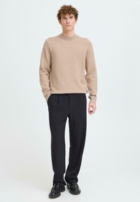 Béžový pletený sveter s okrúhlym výstrihom, v kombinácii s čiernymi plisovanými nohavicami a čiernou obuvou. Jednoduchý dizajn bez vzorov alebo ozdôb.
