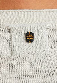 Tissu tricoté gris clair avec un motif texturé. Présente une étiquette noire avec un texte doré "PME Legend" et un petit détail accent doré.