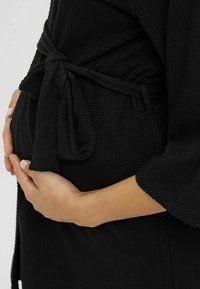 Svart texturerad klänning med en knytning i midjan, bärs över en rundad mage. Synlig hand som försiktigt vilar på tyget.