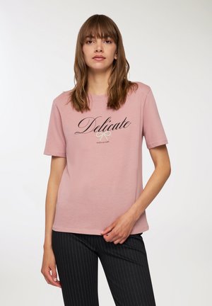 T-shirt rosa a maniche corte con collo a giro, caratterizzato dalla parola "Delicate" in script nero e una grafica a forma di fiocco. Il tessuto appare morbido.