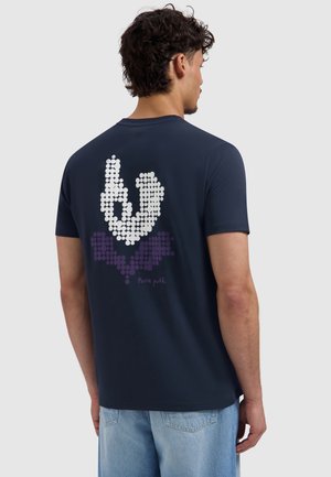 Man met de rug naar de camera, draagt een navy T-shirt met wit en paars stippenpatroon en lichtblauwe spijkerbroek tegen een effen achtergrond.