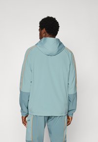 Light blue hooded jacket with orange side accents, featuring a relaxed fit and elastic cuffs. Material appears smooth with a slightly shiny finish.