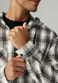 Grau-weißes kariertes Flanellhemd mit Kapuze, das eine geknöpfte Manschette und einen strukturierten Stoff aufweist. Eine Hand ist teilweise sichtbar und greift den Ärmel.