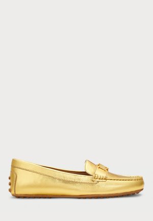Mocassin en cuir doré avec semelle en caoutchouc cloutée et détail de boucle décorative sur le dessus, présenté de profil sur un fond neutre.