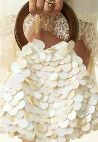 Main avec bague en or tenant une poignée ronde en bois d'un sac recouvert de grands sequins ronds irisés, tissu en dentelle en arrière-plan.