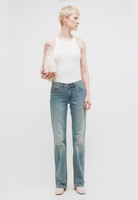 Γυναίκα με κοντά ξανθά μαλλιά που φορεί άσπρο αμάνικο τοπ, ανοιχτό μπλε τζιν, μπεζ παπούτσια, κρατώντας μια μικρή μπεζ τσάντα, κοιτάζοντας προς τα δεξιά.