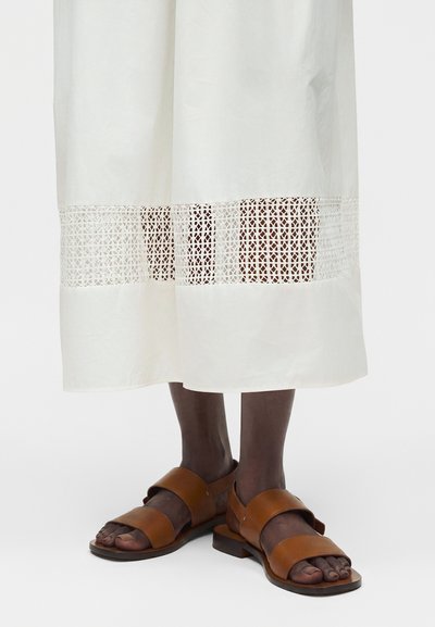 Άτομο που φοράει λευκό φόρεμα με ανοιχτή πλέξη κοντά στο τελείωμα και καφέ δερμάτινα επίπεδα σανδάλια με δύο φαρδιές λουρίδες.